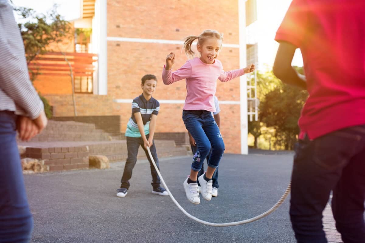 Happy elementary kids playing together with jumping rope outdoor. Children playing skipping rope jumping game and laughing outdoors. Happy cute girl jumping over skipping rope held by her friends.