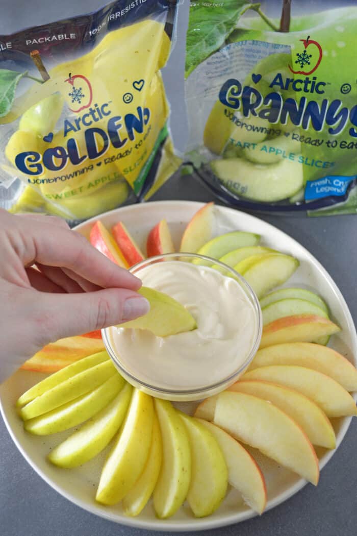 Hand dipping sliced apple into a bowl of caramel yogurt fruit dip with plated with sliced apples. Bags of sliced apples in background.