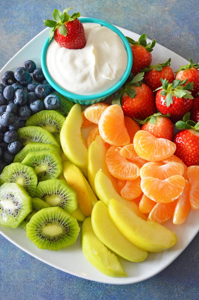 Bowl of cream cheese fruit dip plated with a rainbow of fruit, including blueberries, kiwi, apples, mandarins and strawberries.