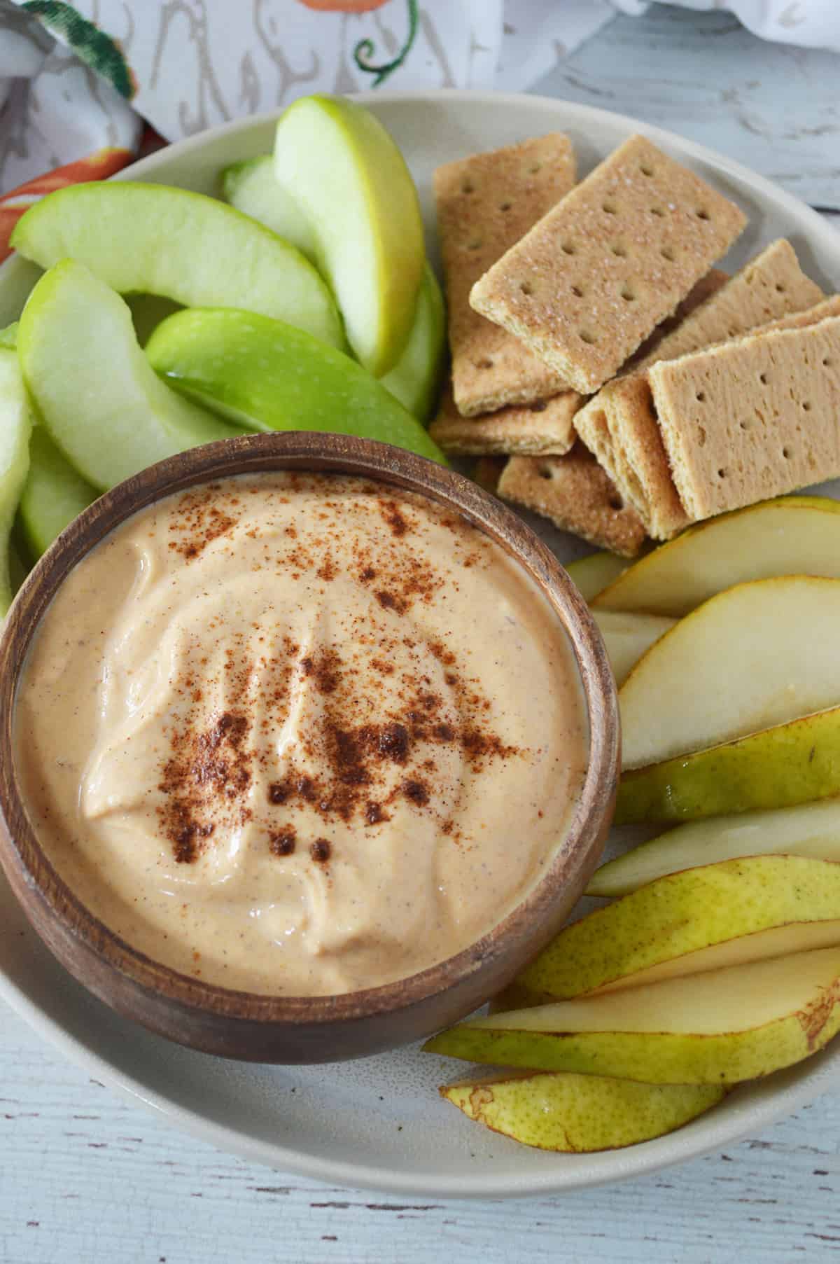 Pumpkin pie dip in bowl on plate with sliced apples, pears and graham crackers.