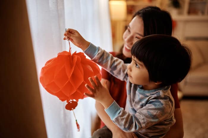 Chinese boy hanging lanterns on window with his mother for Lunar New Year celebration.